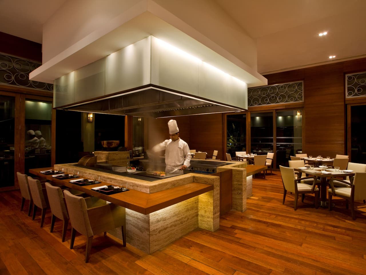 Chef cooking on a teppanyaki grill in a center of the restaurant with the modern wood finish and seating