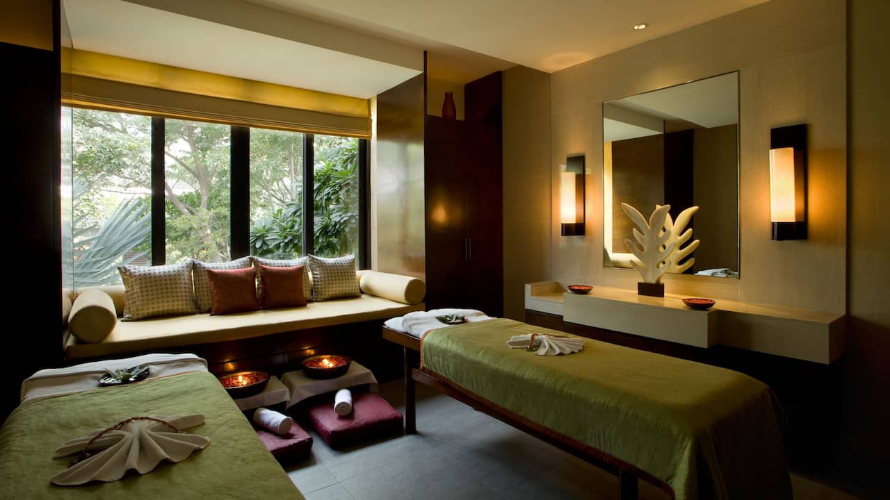 Spa room with setup of massage treatments having 2 separate beds, soft warm light and a large window with lush green view