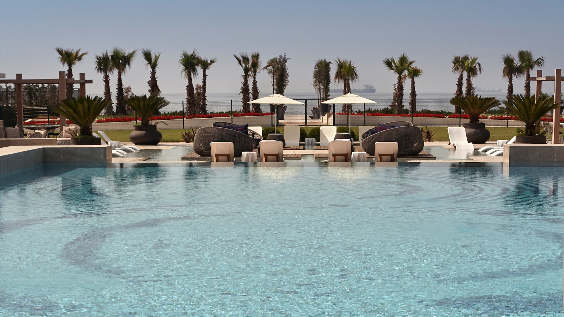 1 of 1 Infinity pool at Hyatt Regency Istanbul Ataköy with submerged lounge chairs, palm trees, and a sea view in the background.