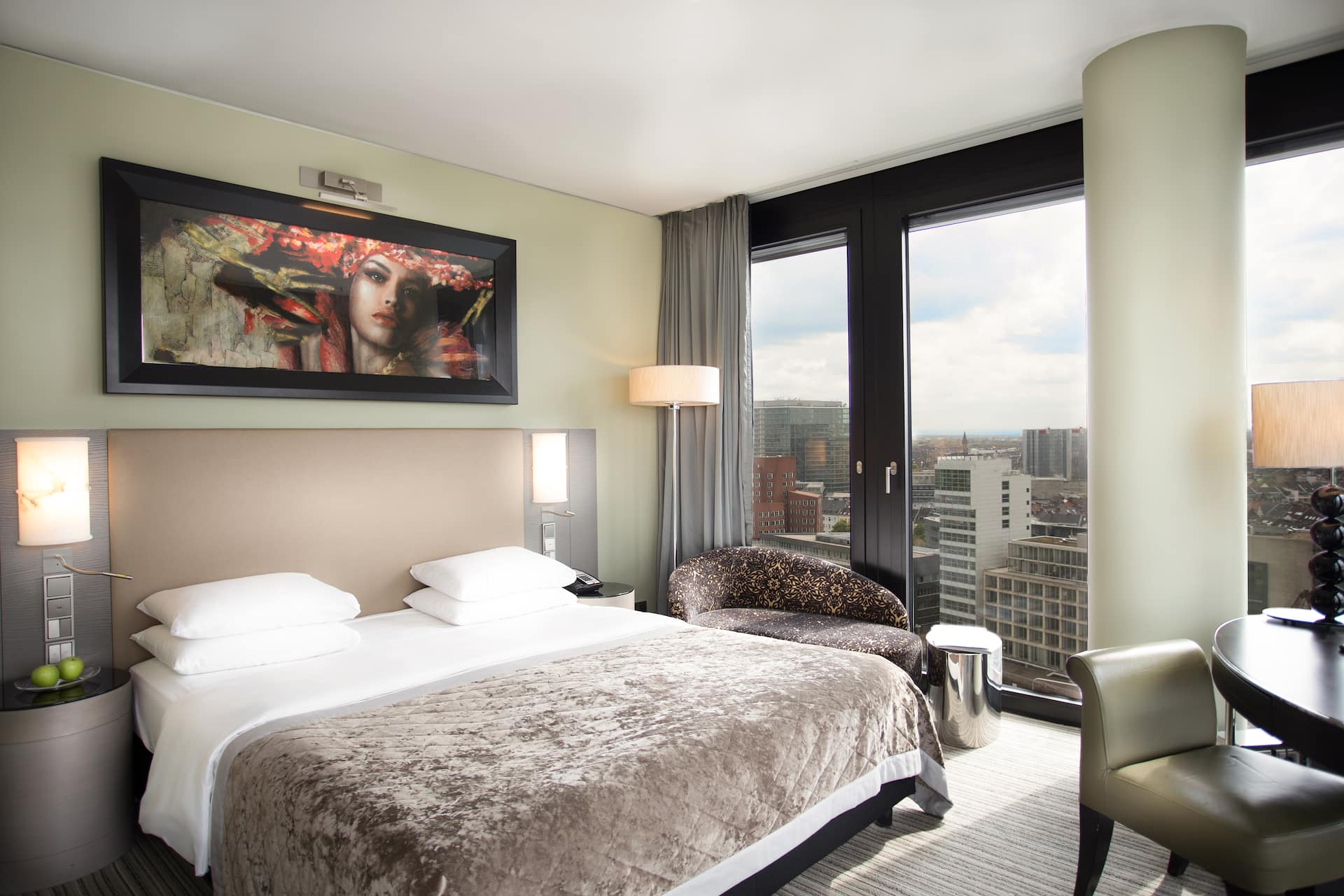 King Deluxe Room with king-size bed at the Hyatt Regency Dusseldorf with floor-to-ceiling windows, a small couch, warm earth tones and a desk for working