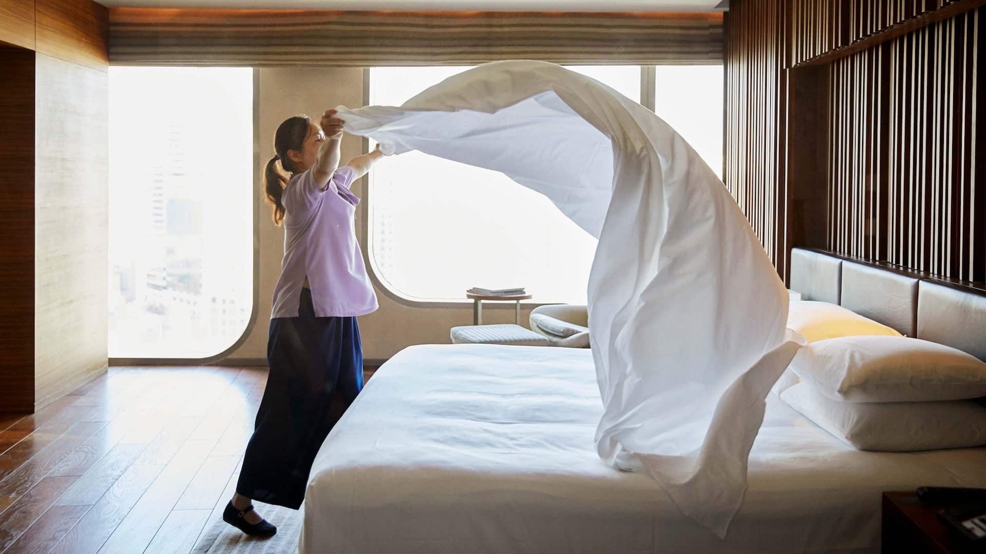 A hotel worker billows out a fresh sheet over a guest bed.