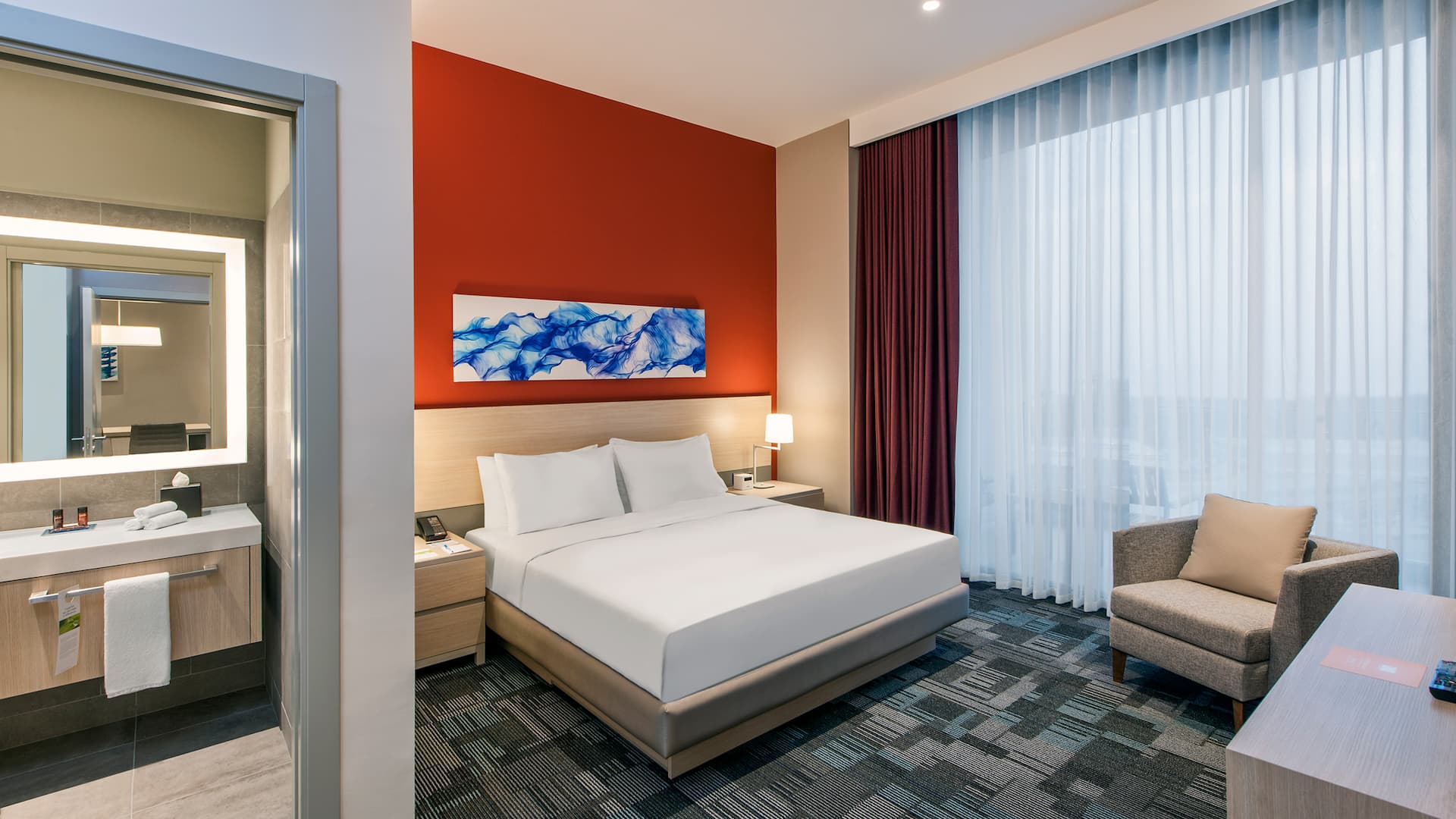 One-bedroom suite in Gebze with a king bed, soft chair, red accent wall, blue artwork, closed curtains, and partial bathroom view at Hyatt House Gebze, perfect for a comfortable stay.
