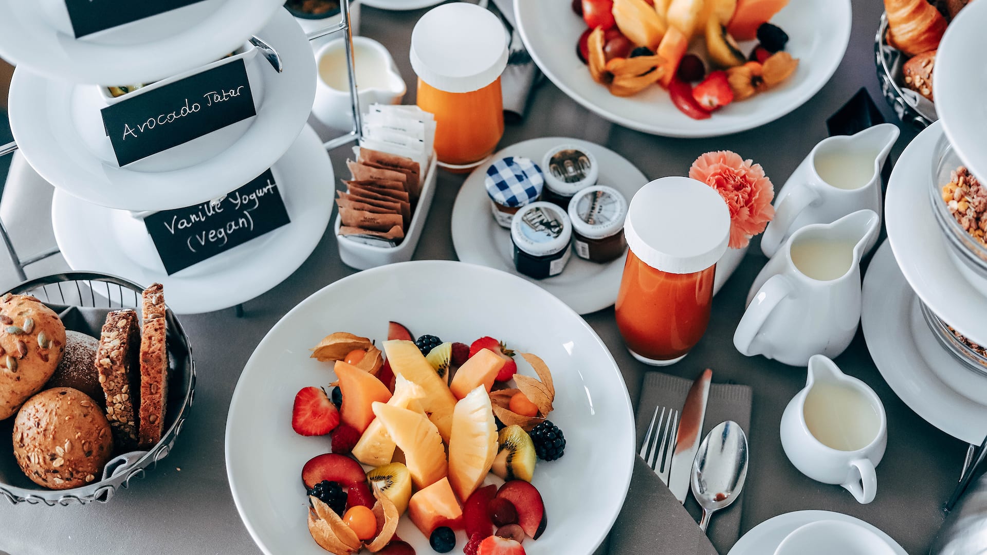 Roomservice Frühstück mit einer Auswahl an frischem Obst, Brötchen, Marmeladen, Müsli, Joghurt, Milch und Avocado.
