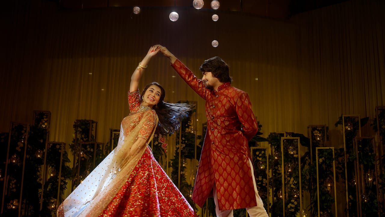 Couple dancing in traditional attire smiling to each other with wedding setup in backdrop having overall warm ambience