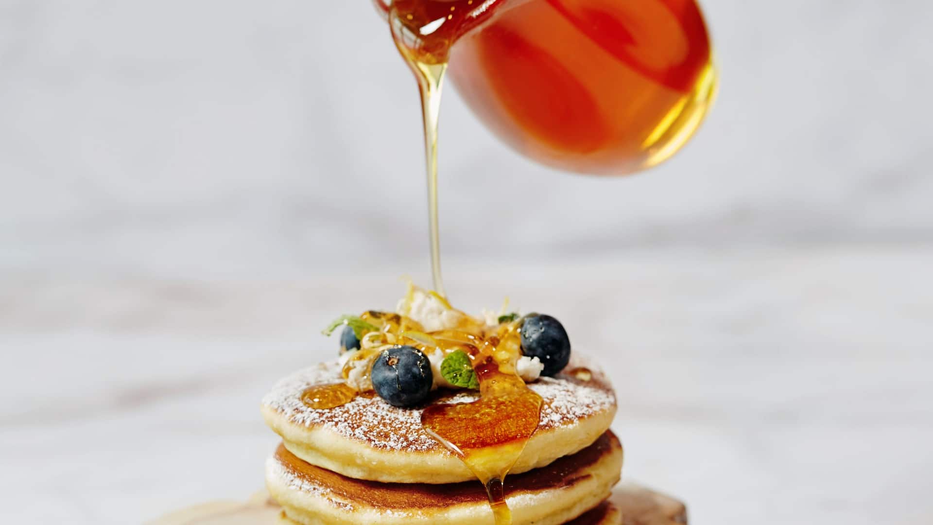 label.xOfY A stack of fluffy pancakes topped with blueberries, honey drizzle, and garnish, with golden syrup being poured over them.