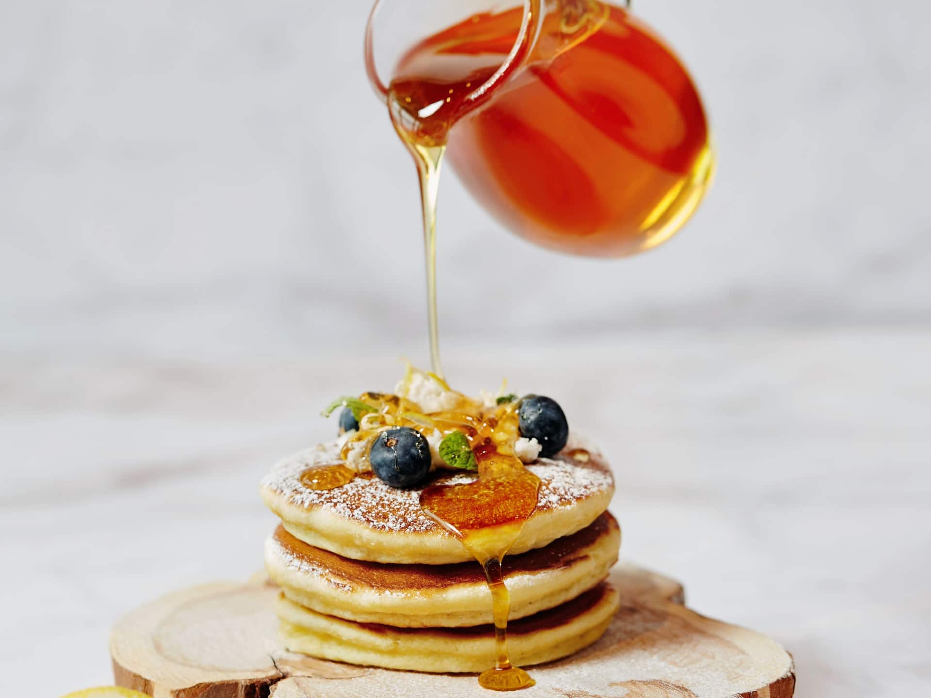 A stack of fluffy pancakes topped with blueberries, honey drizzle, and garnish, with golden syrup being poured over them.