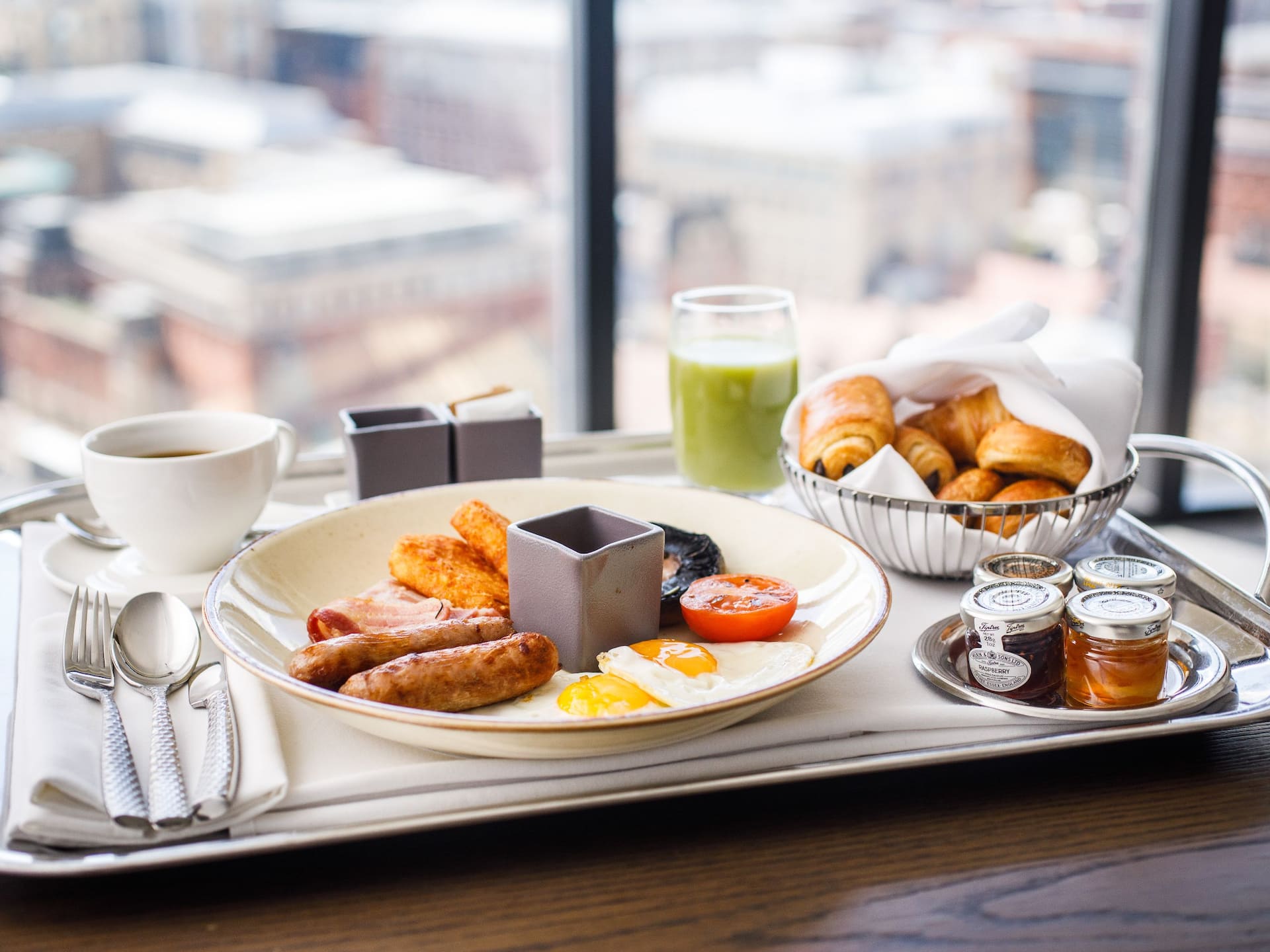 Tray on a wooden table with a classic breakfast composed of croissants, pain au chocolat, an English breakfast with eggs, beans, sausages and a coffee with a view of Birmingham.