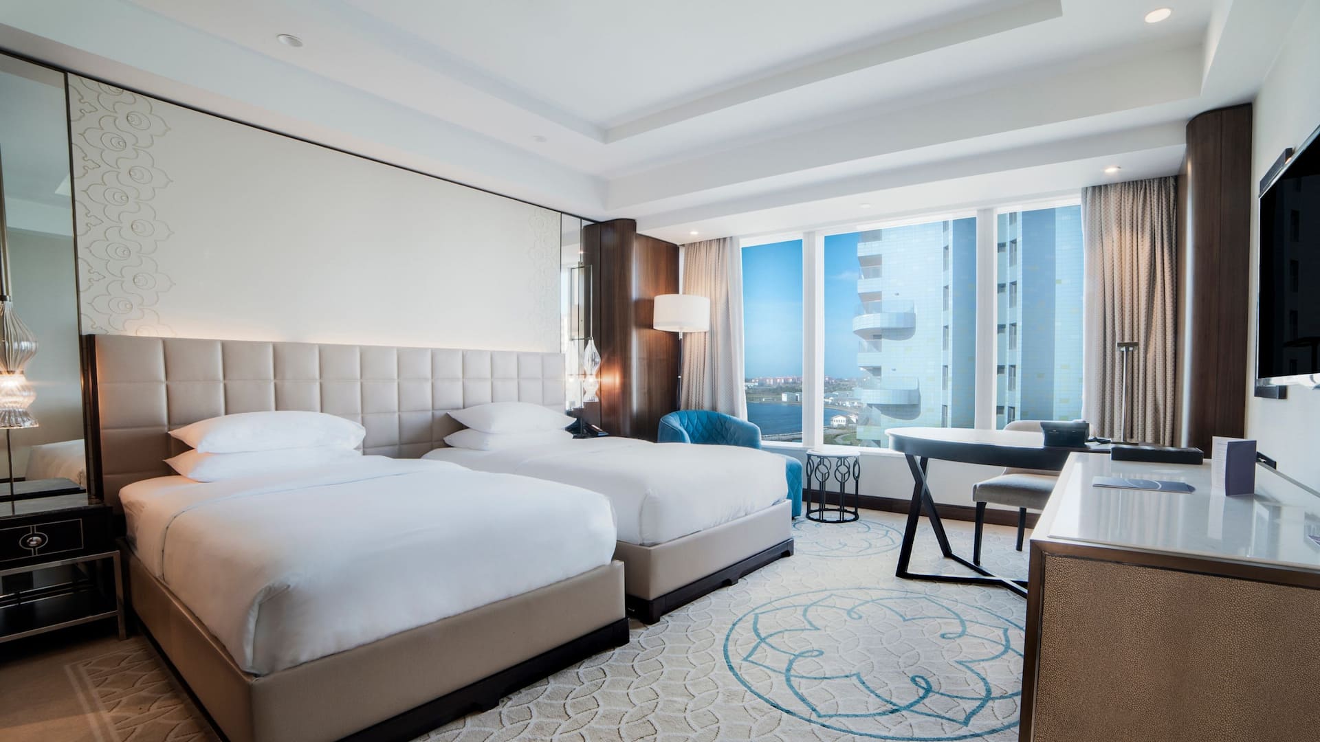 2 Twin Beds at Hyatt Regency Istanbul Ataköy with a tufted beige headboard, plush blue armchair, and floor-to-ceiling windows offering city views.