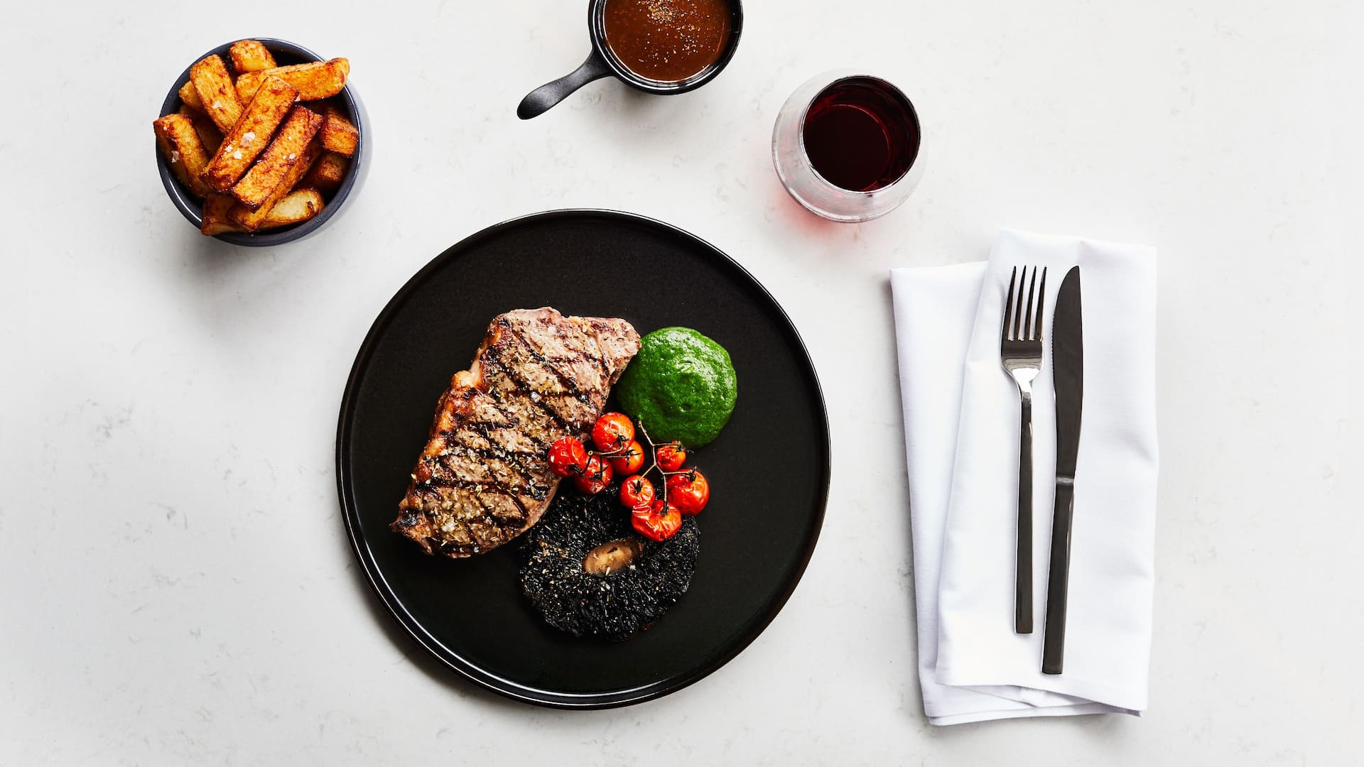 Cooked steak with mushroom, tomatoes and spinach on a black plate on a white marble table with chips, glass of red wine and cutlery
