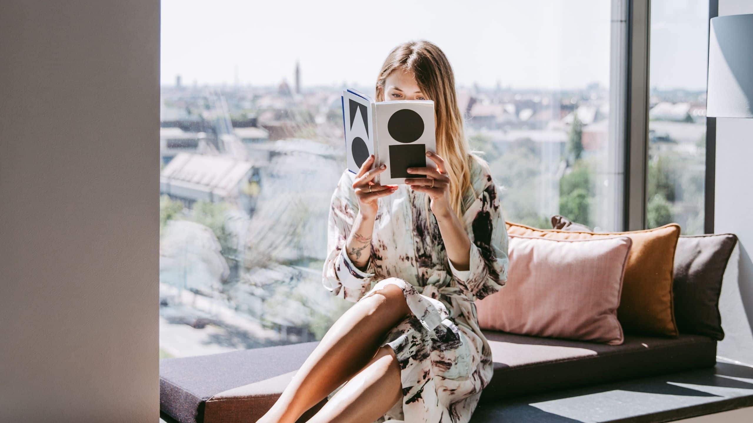 A woman sits by a large window, reading a book in the modern, cozy Andaz Munich suite with a city view