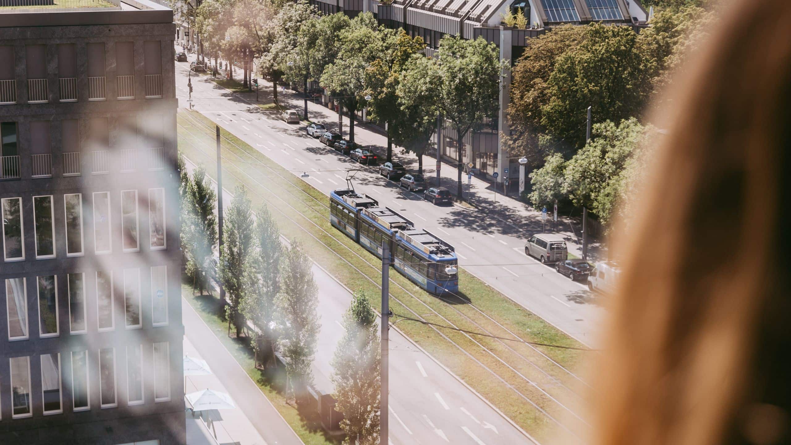 A woman looks out of an upper floor window at Andaz Munich hotel, viewing a tram passing through the city streets below