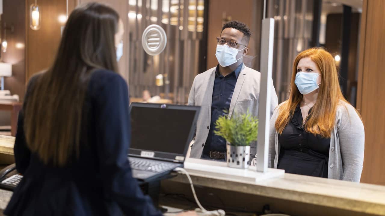 Front Desk Agent Behind Safety Glass w Face Mask Checking In Guests w Face Masks