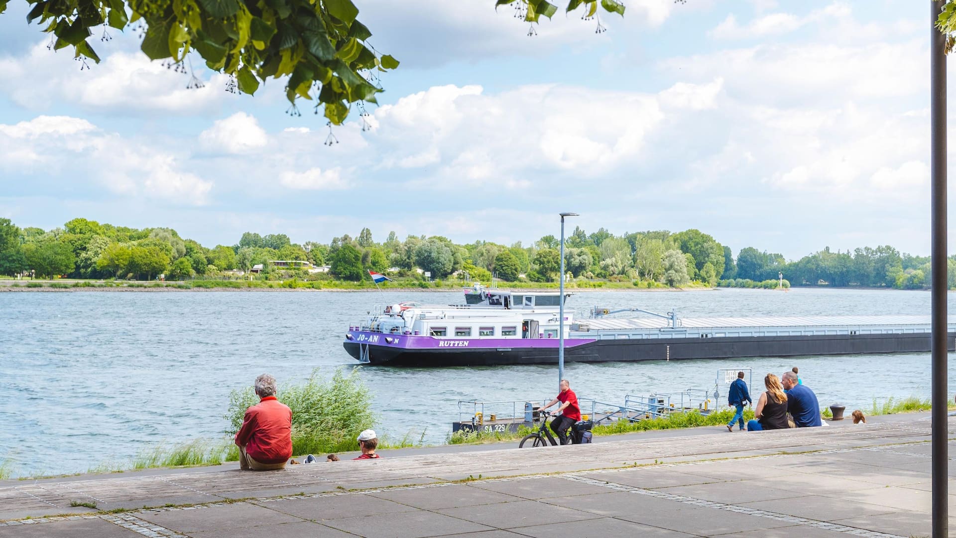 People sitting on Rhine piazza in front of Hyatt Regency Mainz watching ships pass by