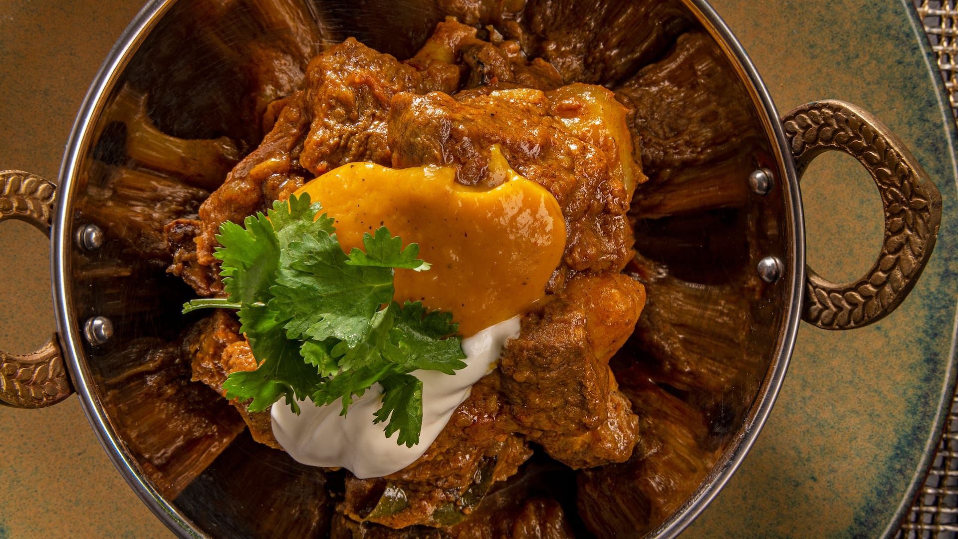 Close-up of a flavorful Cape Malay curry served in a traditional brass-handled bowl, topped with creamy yogurt, tangy mango chutney, and fresh cilantro — a signature dish at a Cape Town hotel restaurant, ideal for food lovers and culinary tourism.