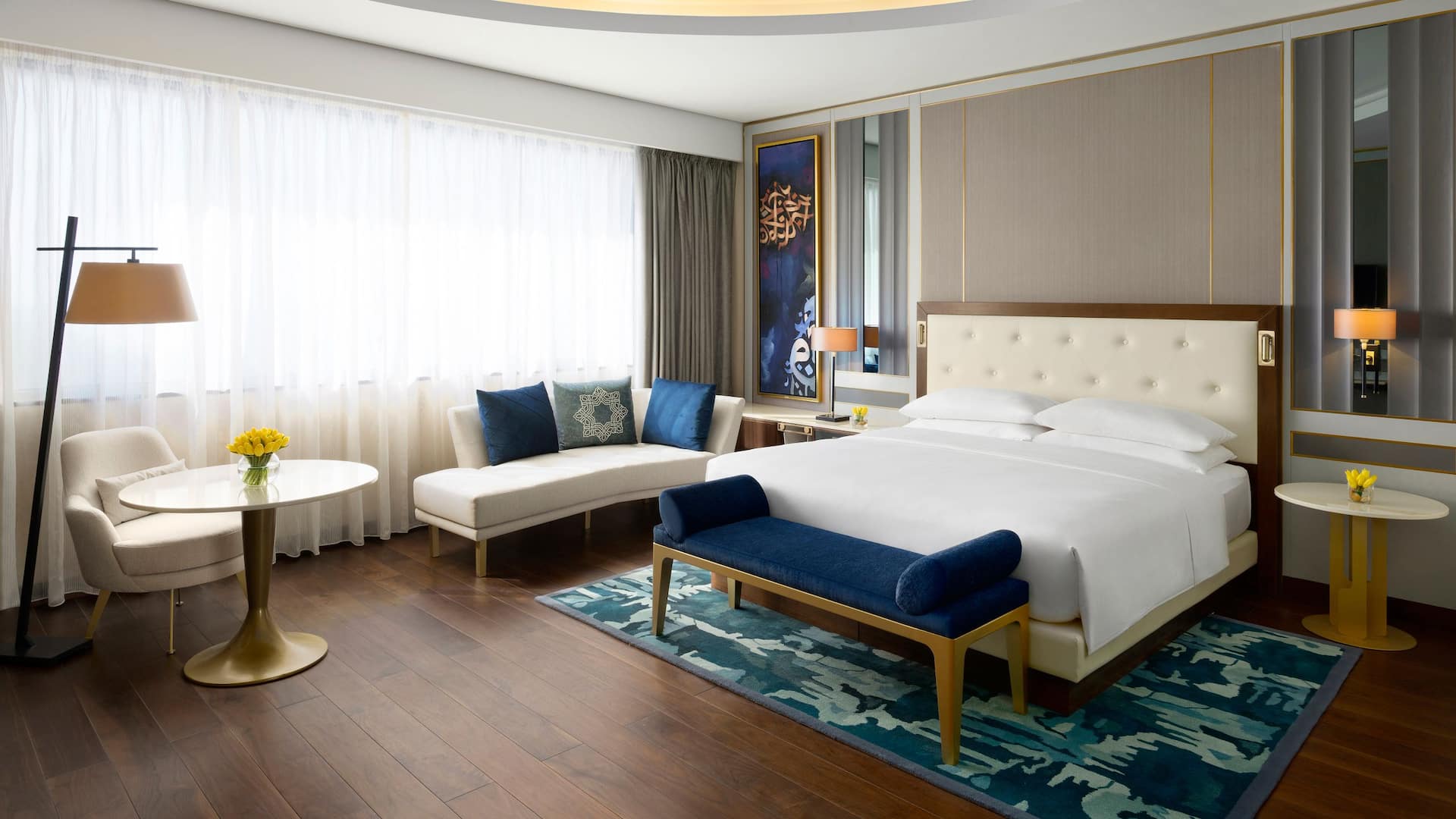 4/6 Elegant guest room at Grand Hyatt Al Khobar featuring a plush king size bed with a tufted headboard, a stylish seating area with a white chaise lounge, and a gold accented round table, The room is adorned with modern Arabian inspired decor