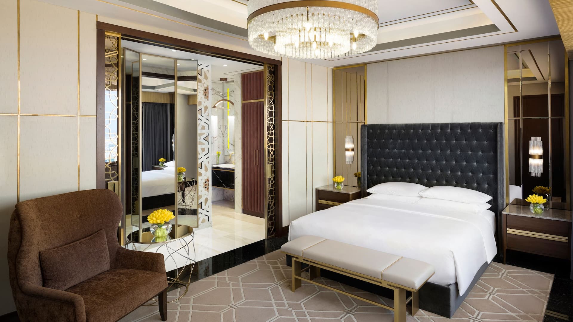 1 of 8 Elegant hotel room at Grand Hyatt Al Khobar featuring a king sized bed with a tufted black headboard, a plush brown armchair, and a luxurious crystal chandelier, The room includes gold accented decor, mirrored panels
