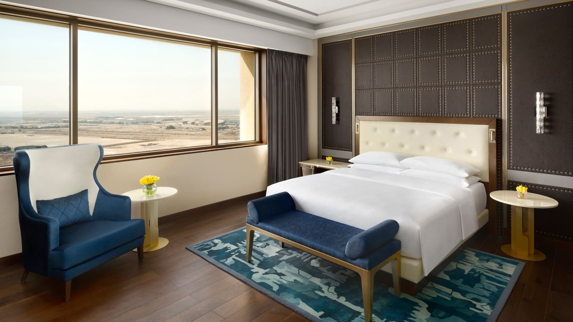Luxurious hotel room at Grand Hyatt Al Khobar featuring a king sized bed with a tufted cream headboard, a stylish blue and white lounge chair, a plush blue bench, and elegant gold accented side tables