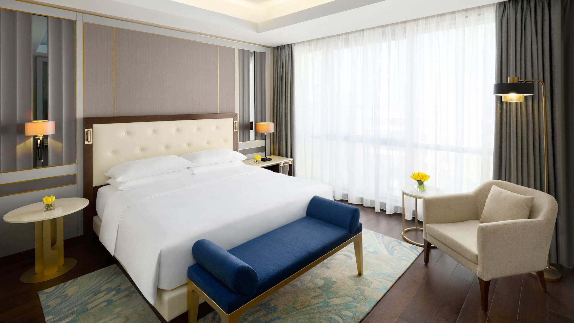 1/6 Elegant and modern hotel room at Grand Hyatt Al Khobar, featuring a king sized bed with a tufted cream headboard, a plush blue bench, and gold accented decor, The room is bathed in natural light from large windows with sheer white curtains