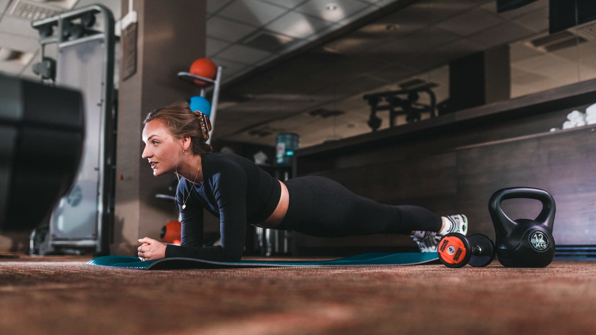 A woman performing a plank exercise on a mat in the Hyatt Place Amsterdam Airport gym, with kettlebells and dumbbells beside her.