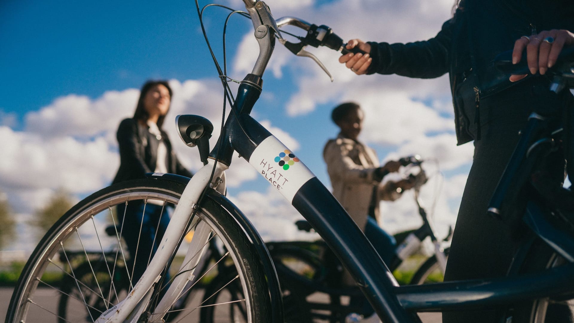 A close-up of a bike with a "Hyatt Place" logo, with two people riding bicycles in the background under a blue sky with clouds.