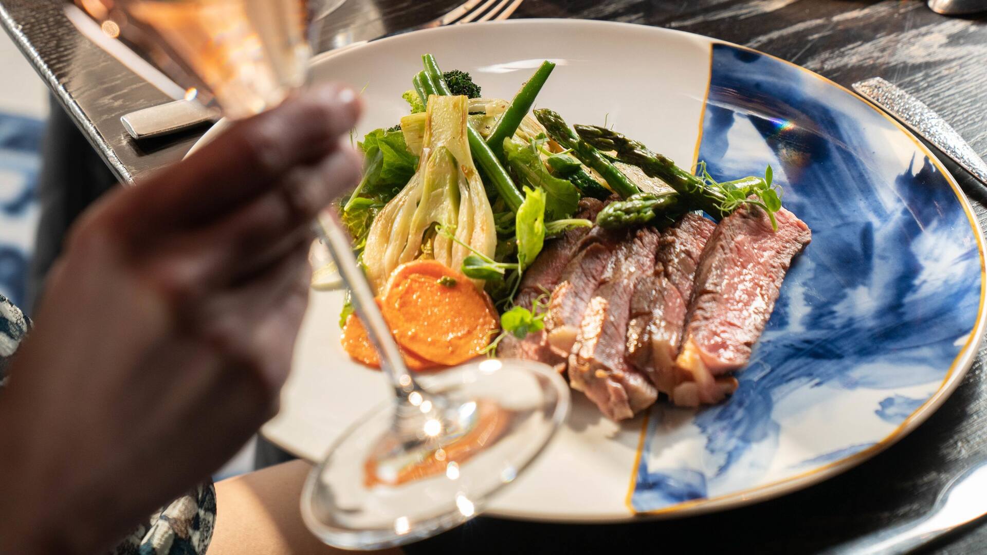 label.xOfY A gourmet plate featuring perfectly seared steak, sautéed vegetables, and a vibrant presentation, with a hand holding a glass of champagne in the foreground.