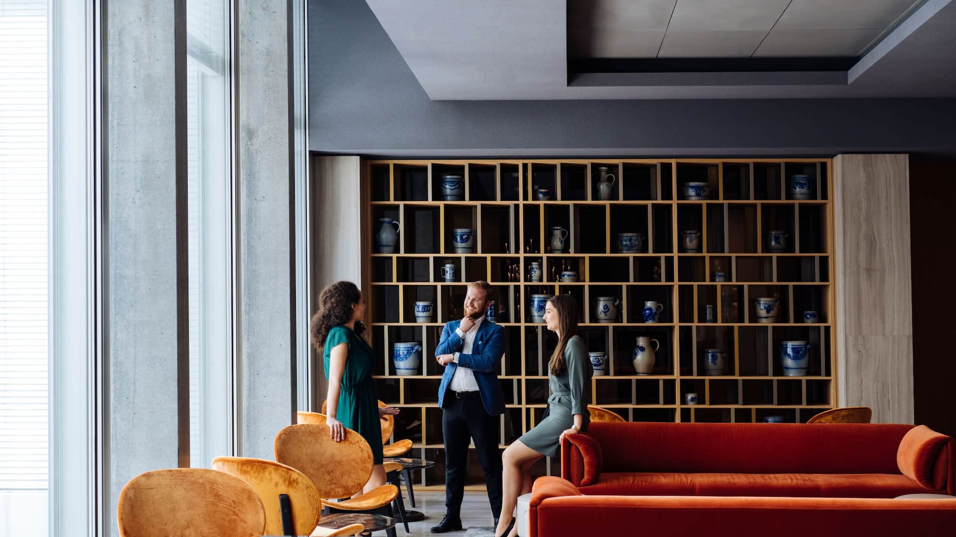 Three business professionals casually conversing in a modern, stylish lounge at Hyatt Regency Zurich Airport The Circle, with a wooden shelf featuring blue and white ceramic decor as the backdrop.
