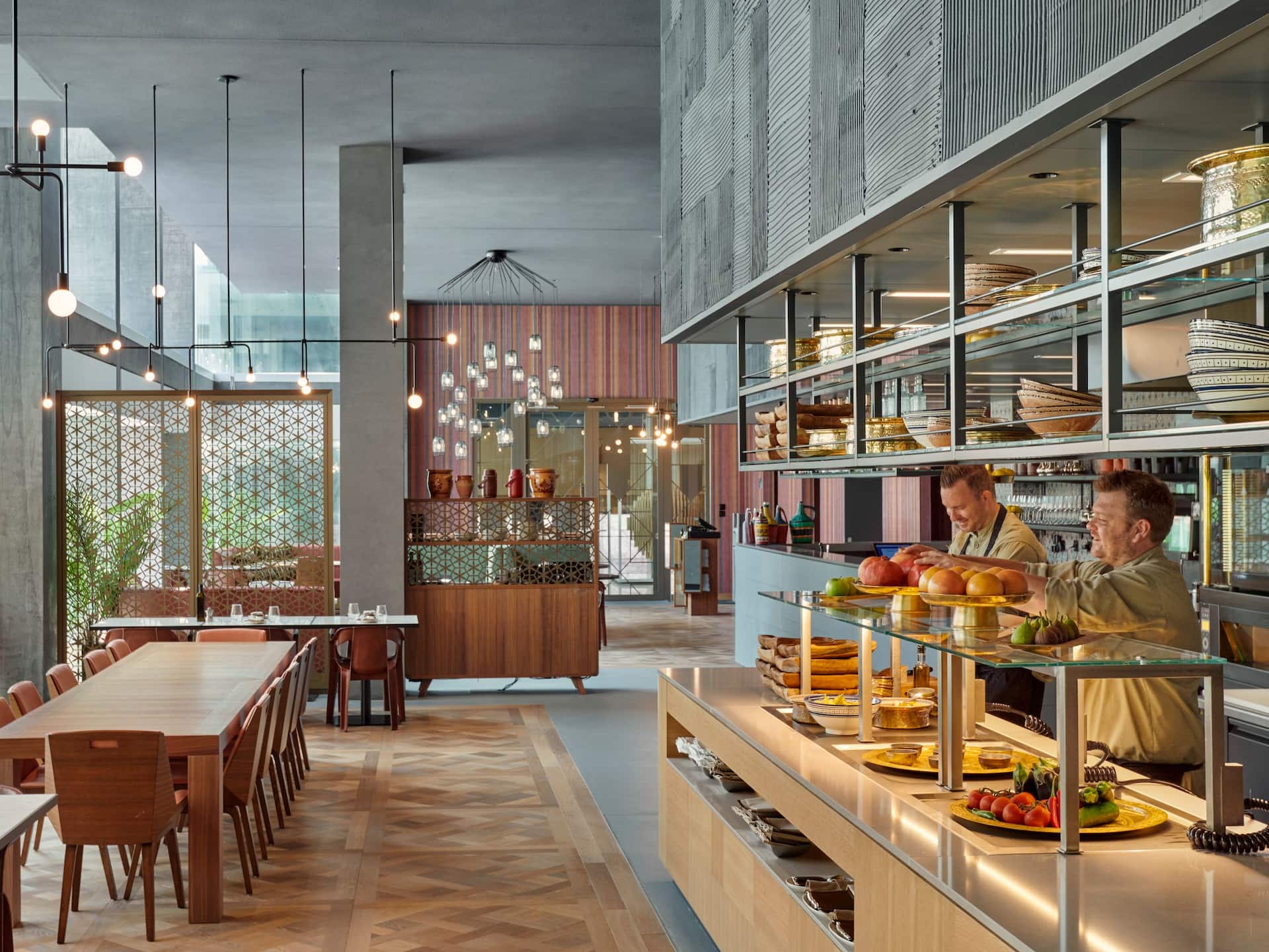 Interior view of Babel Restaurant, a Lebanese dining venue featuring modern decor with wooden tables and chairs and a bright, open atmosphere. The image showcases a serving counter with a variety of dishes and two staff members preparing food.
