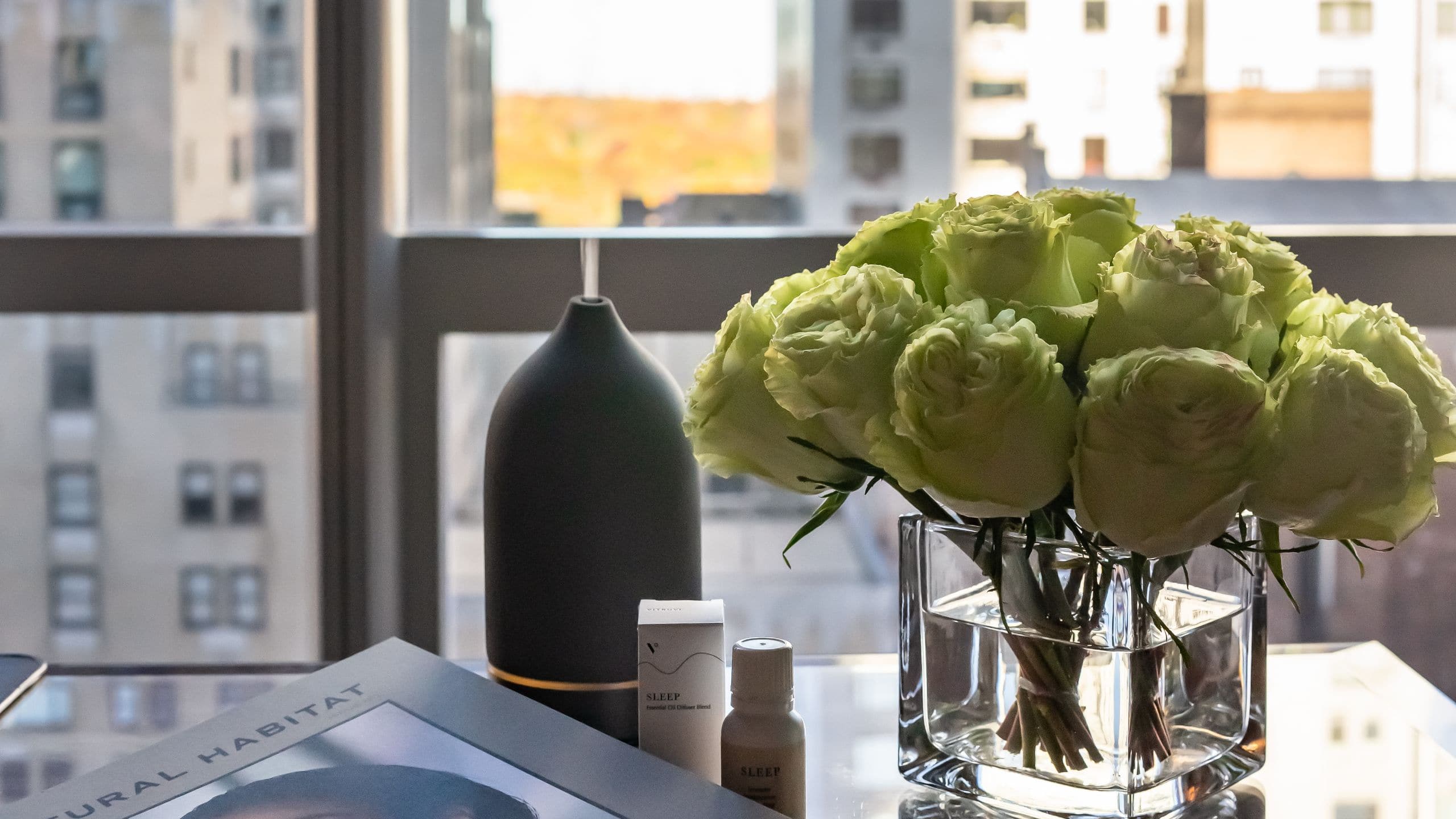 Some flowers in a vase and book on a table in front of large windows with a city view