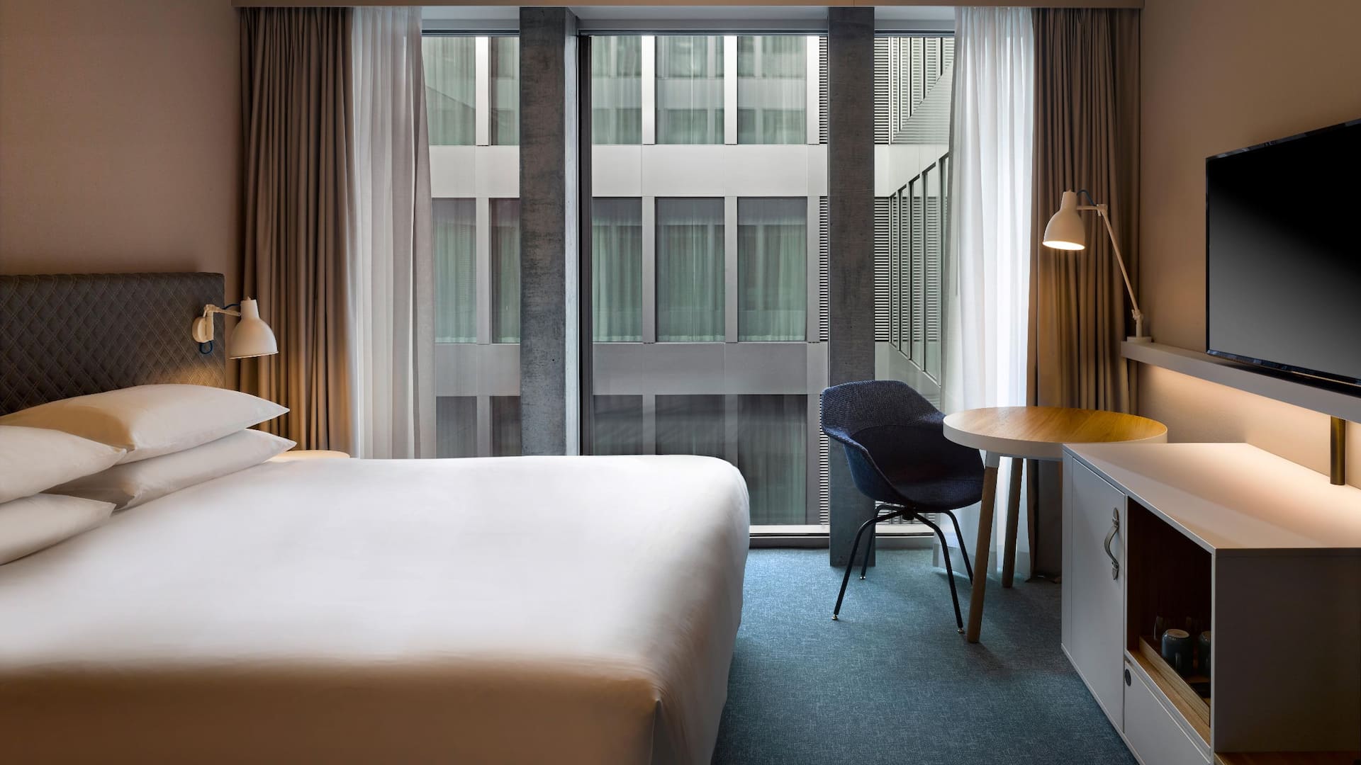 A hotel room in Zurich with one king bed, floor-to-ceiling windows, a cosy work desk and an interior view of the Circle.