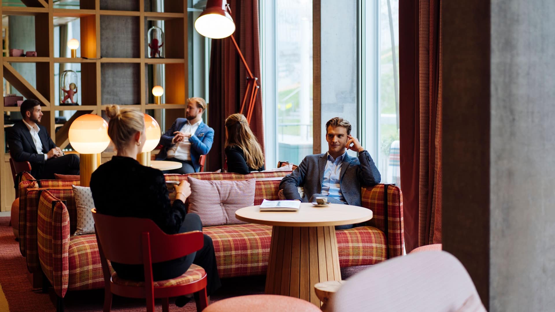 1 of 2 A cozy restaurant lounge at a Zurich hotel with people engaged in conversations, featuring warm lighting, plaid seating, and a modern ambiance.