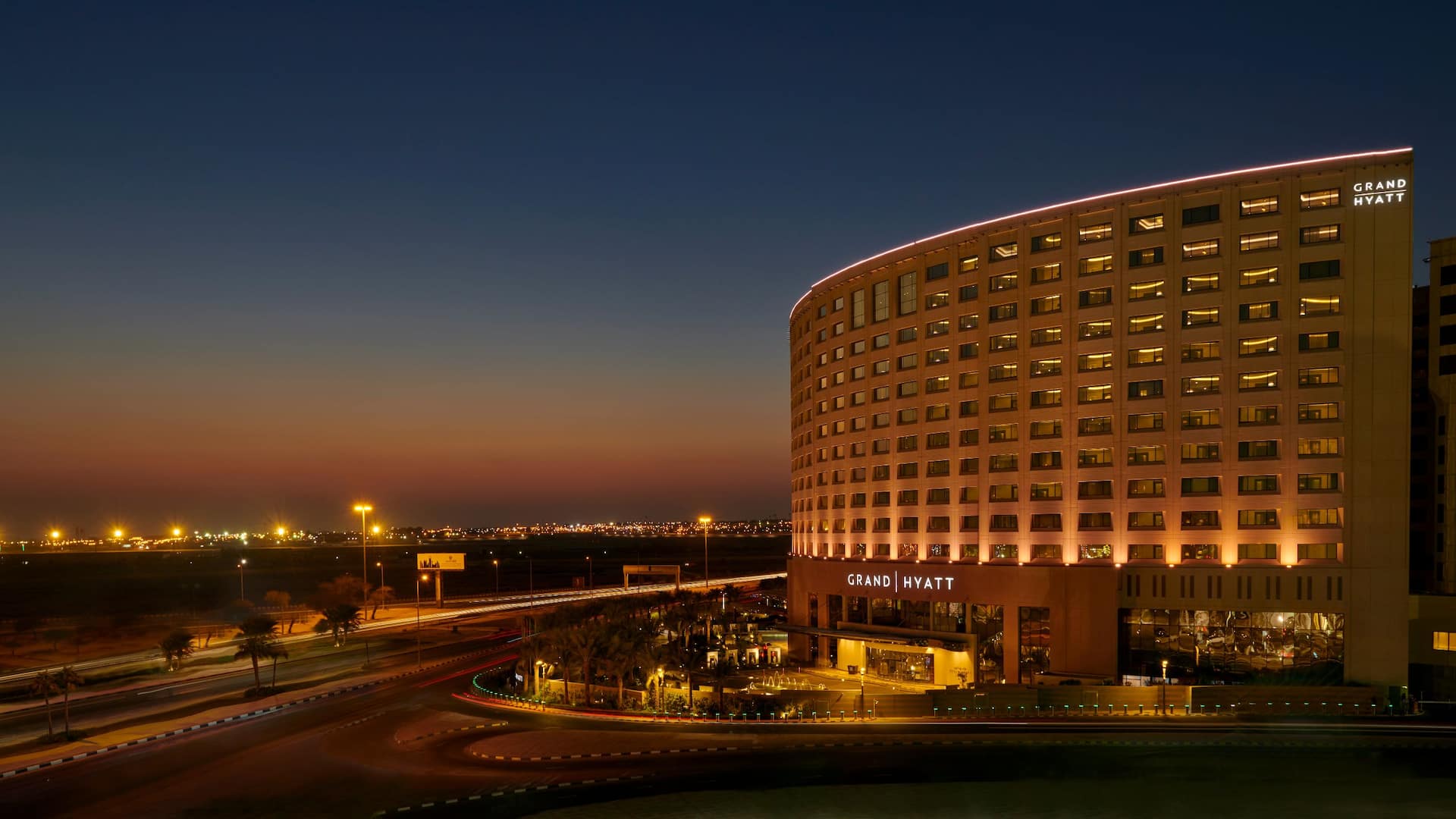 Nighttime view of the Grand Hyatt Al Khobar hotel, a modern curved building illuminated by warm golden lights, The hotel name is displayed on the facade, and the entrance is brightly lit