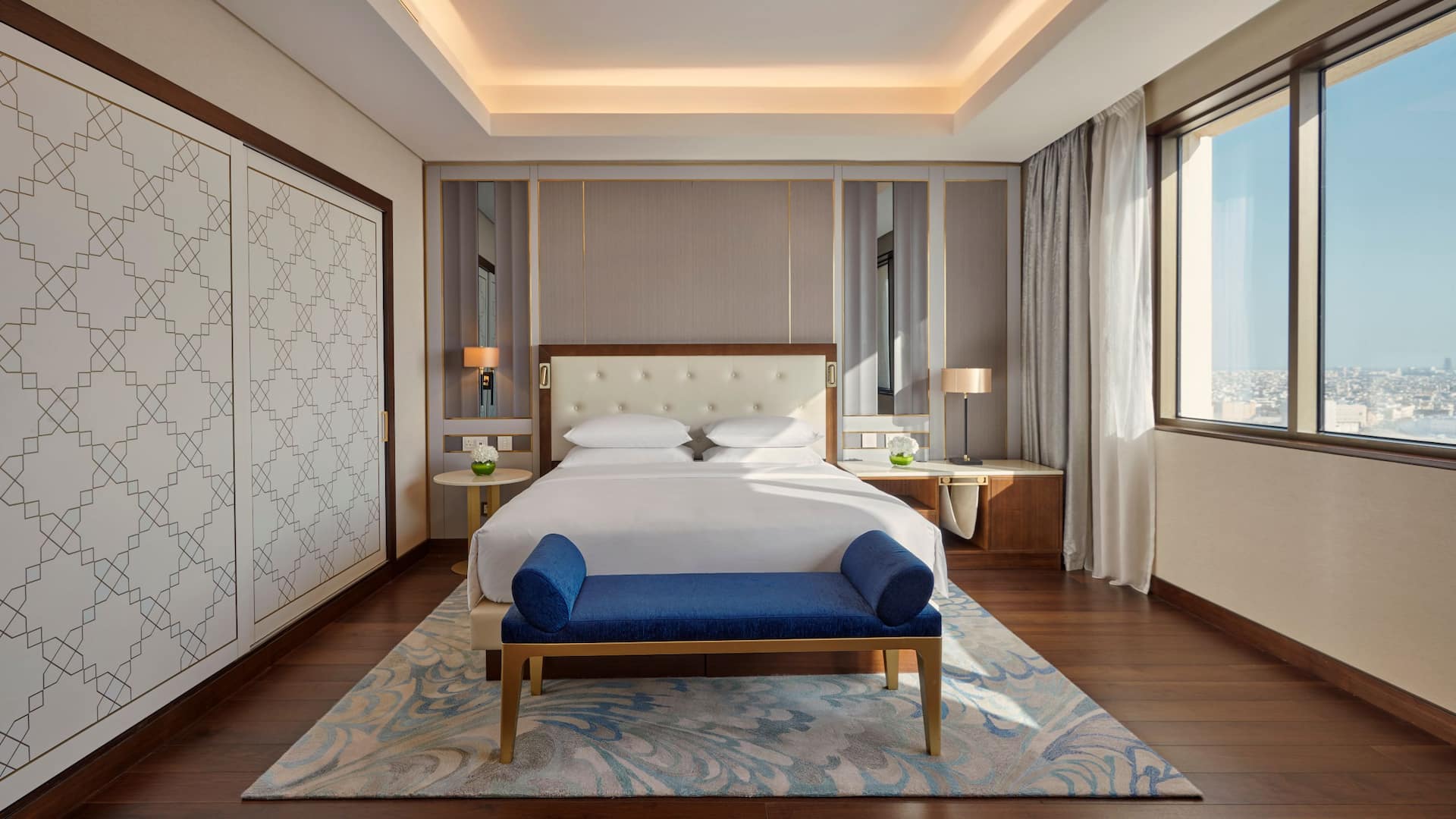 1/5 Modern hotel room at Grand Hyatt Al Khobar featuring a king sized bed with a tufted cream headboard, a plush blue bench, and warm wooden flooring, The room is illuminated by natural light from large windows offering a stunning city view