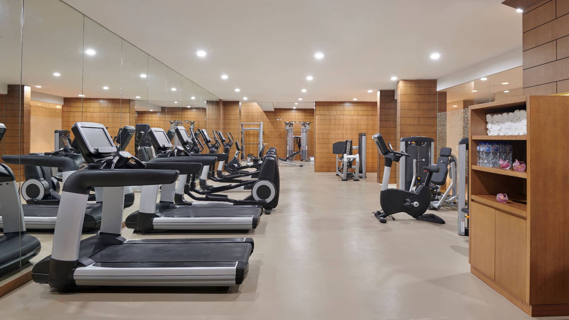 A spacious and well lit fitness center with modern exercise equipment, including multiple treadmills, elliptical machines, and strength training machines, The gym features a neutral toned design with beige flooring, warm wooden panel walls