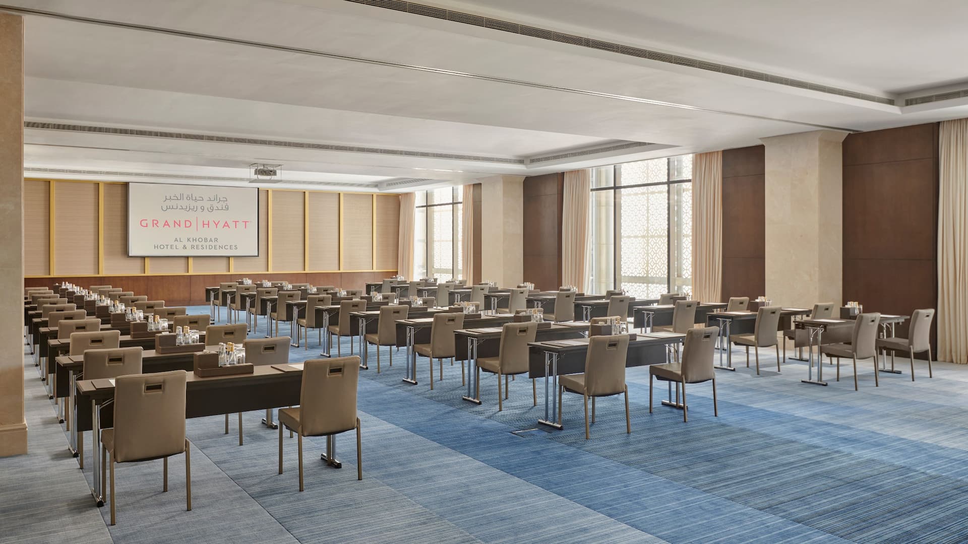 A spacious and modern conference room set up for a business meeting or seminar, Rows of sleek dark brown tables and beige chairs are arranged in a classroom style layout
