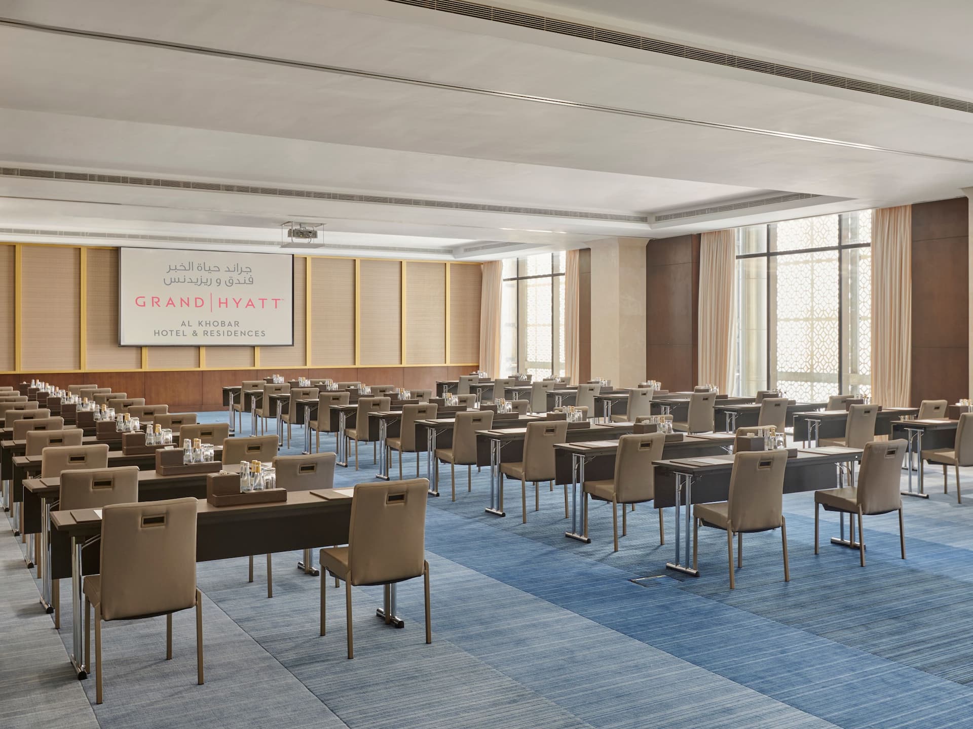 A spacious and modern conference room set up for a business meeting or seminar, Rows of sleek dark brown tables and beige chairs are arranged in a classroom style layout
