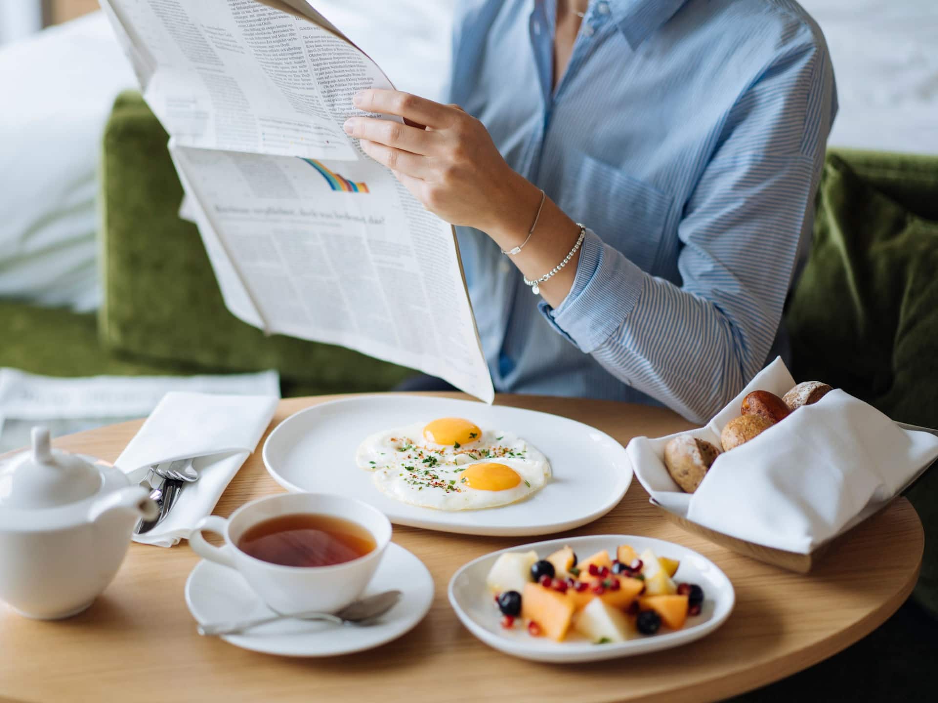 Room service setup featuring a wooden table with a breakfast spread, including fried eggs, fresh fruit, bread rolls, and tea. A person reads a newspaper in the background, creating a relaxed and cozy atmosphere.