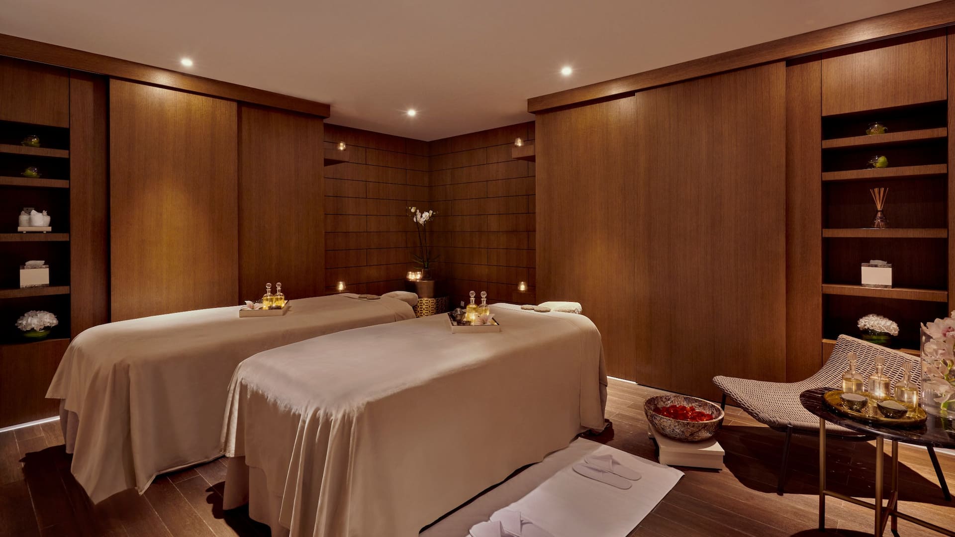 A luxurious spa room featuring two massage tables draped in beige linens, set in a warmly lit ambiance, The space has rich wooden panel walls, soft recessed lighting, and decorative elements such as an elegant orchid in a vase
