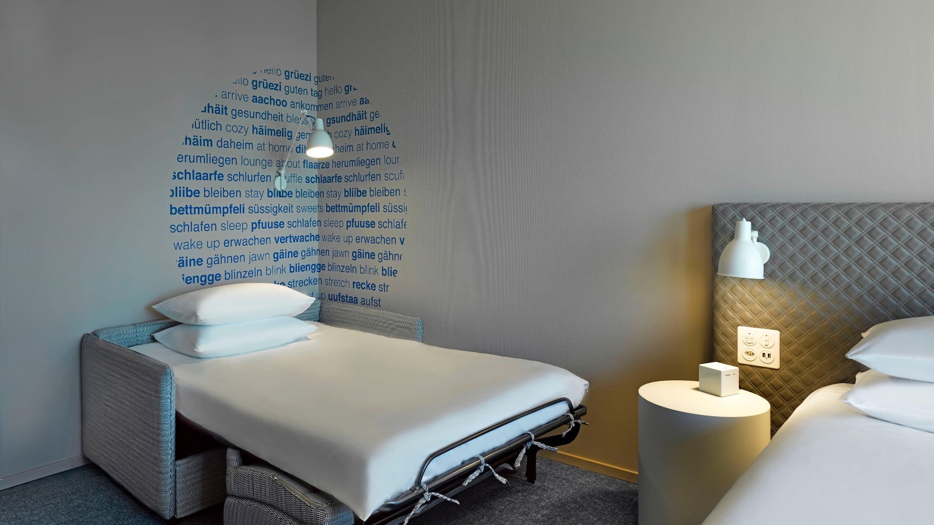 A hotel room in Zurich featuring a king bed and a cozy corner with a sleeper sofa, a work desk and floor-to-ceiling windows with iews over the airport or park.