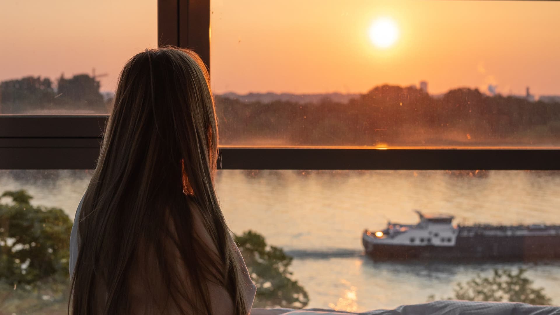 Woman sits in bed facing the large window and enjoying sunset over the Rhine river in room of Hyatt Regency Mainz