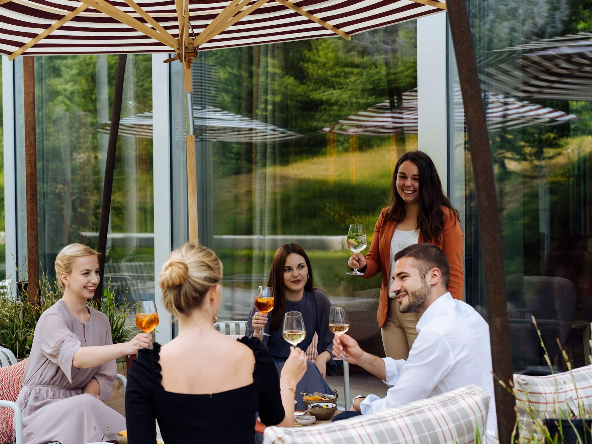 A group of friends enjoying drinks on an outdoor terrace with modern seating, surrounded by greenery and natural light at a Zurich Airport hotel.