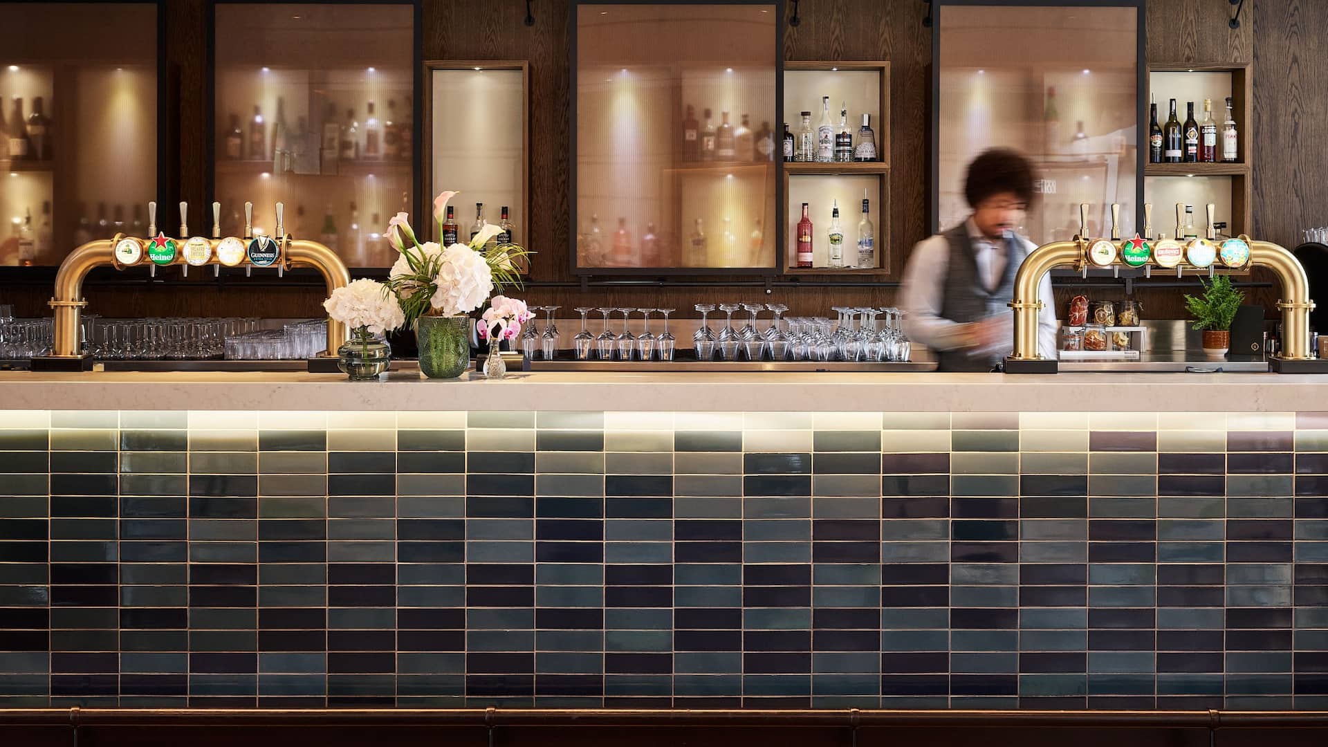 The Graduate Bar tiled front with brass beer pumps and flowers on the counter and a bar tender preparing a drink