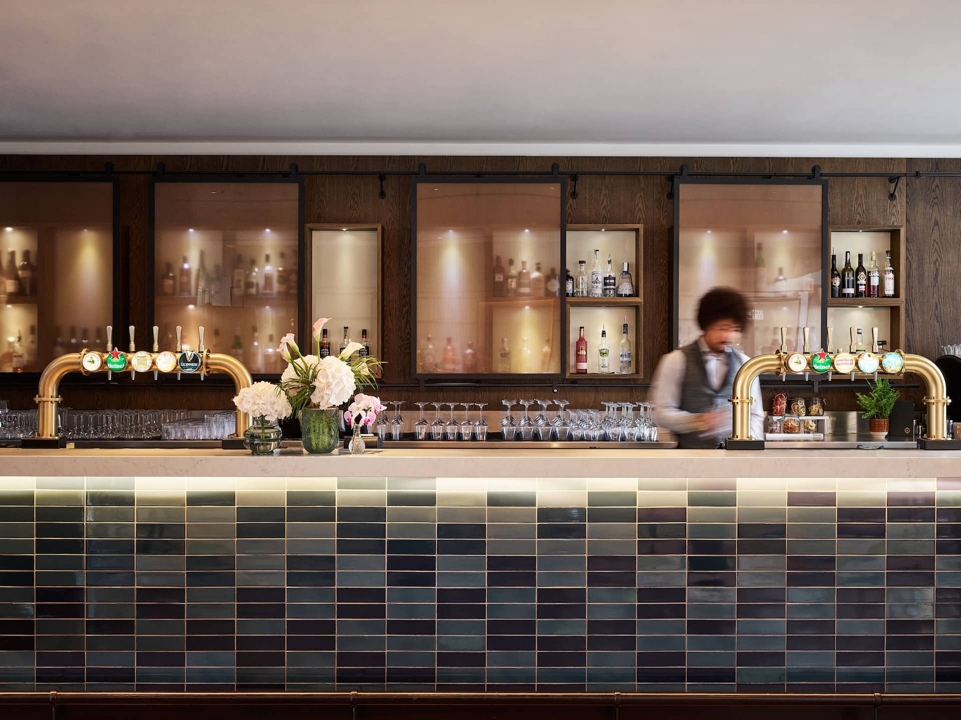The Graduate Bar tiled front with brass beer pumps and flowers on the counter and a bar tender preparing a drink
