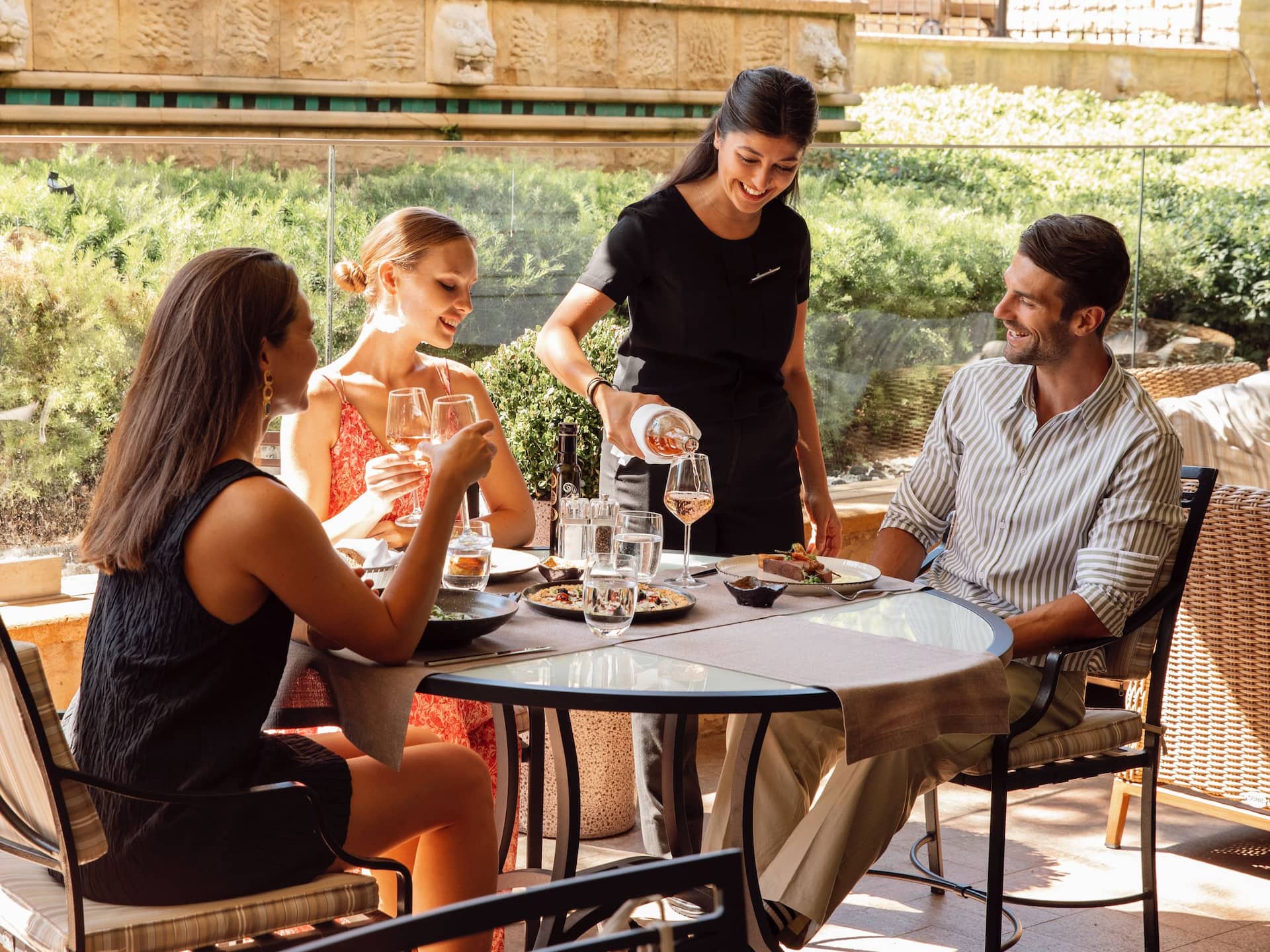 34 Restaurant green surrounded terrace at Grand Hyatt Istanbul with 3 guests enjoying lunch, server pouring wine, starters on the table in a pleasant weather.