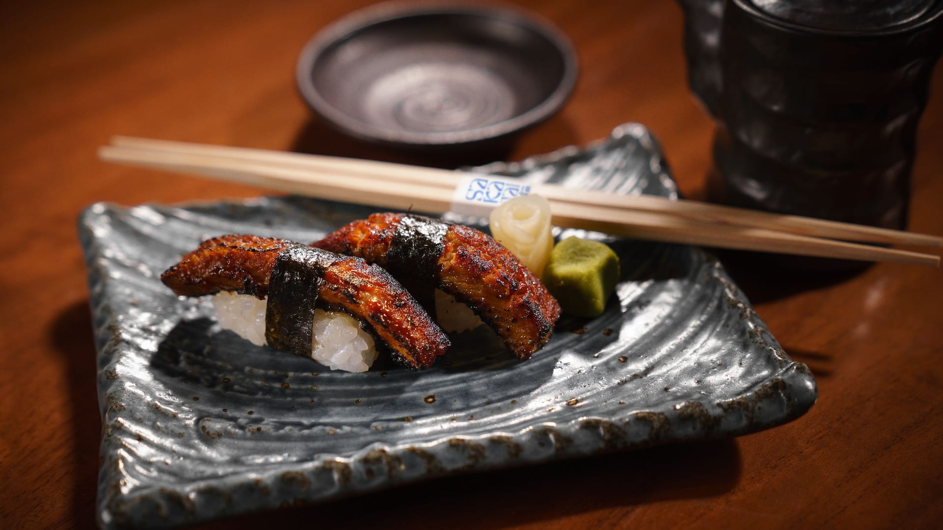 A close up of two pieces of unagi nigiri sushi, featuring glazed and grilled eel on a bed of sushi rice, secured with a strip of nori seaweed at Sakana House Multi Asian restaurant