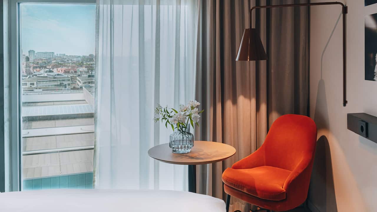 Armchair by the curtains lit up by fashionable lamp and flowers on table 
