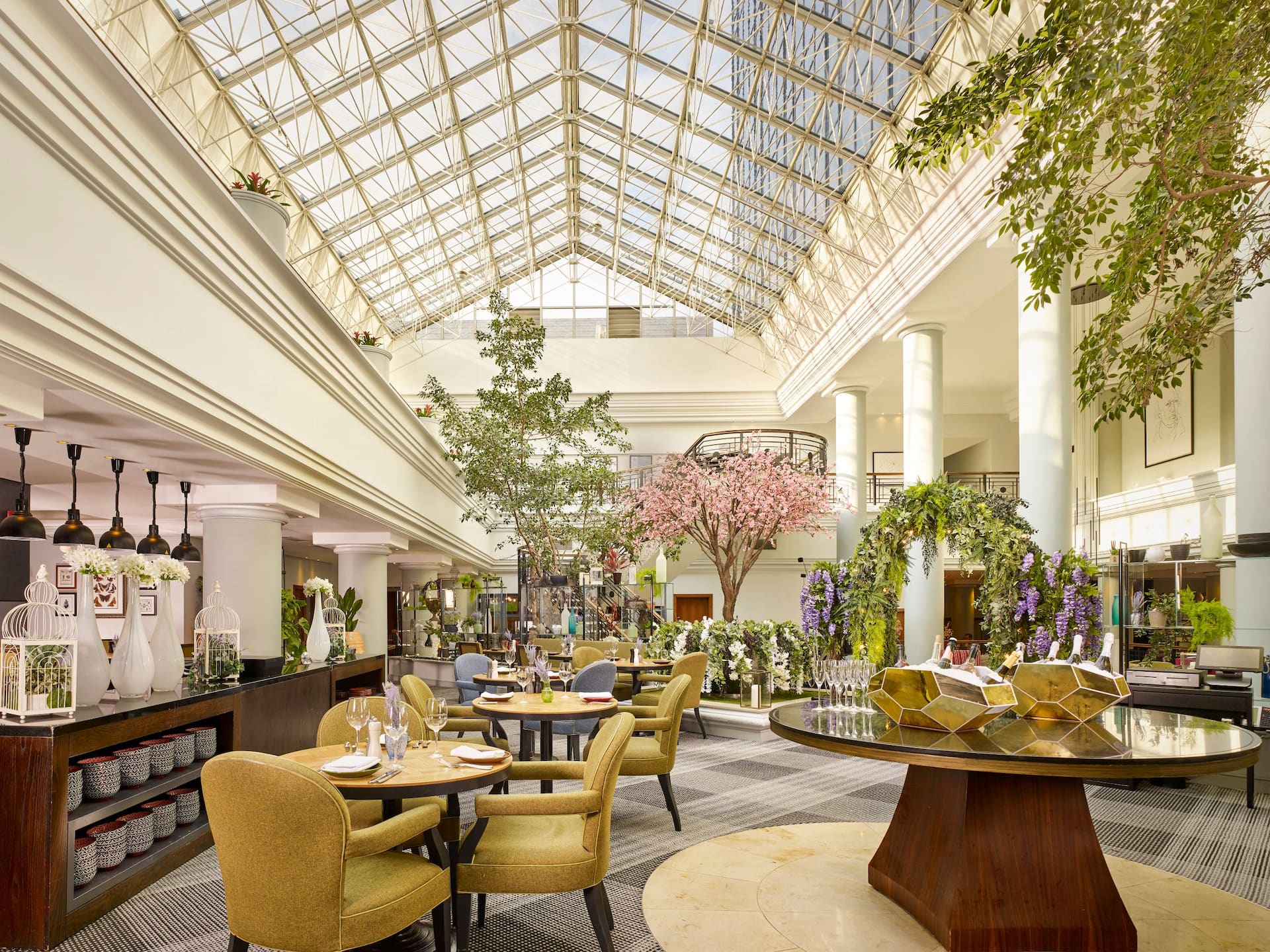 Rond high table presenting bottles of Champagne at Aria Restaurant in Birmingham, surrounded by tables, chairs and lush greenery under the atrium.