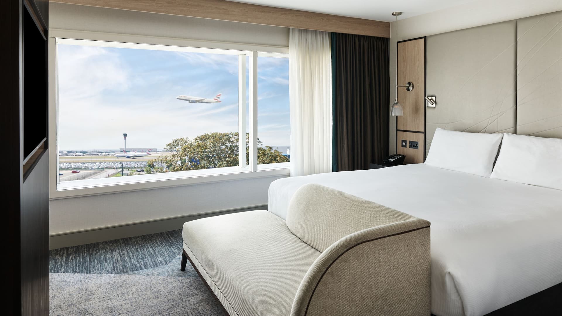 Deluxe room with king bed at London Heathrow Airport hotel, featuring stylish decor, armchair, and scenic view.