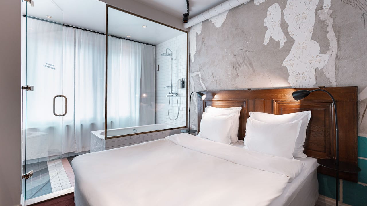 Story Hotel Riddargatan queen deluxe bedroom viewed from entrance, showing bed and glass walls separating room and bathroom with bathtub