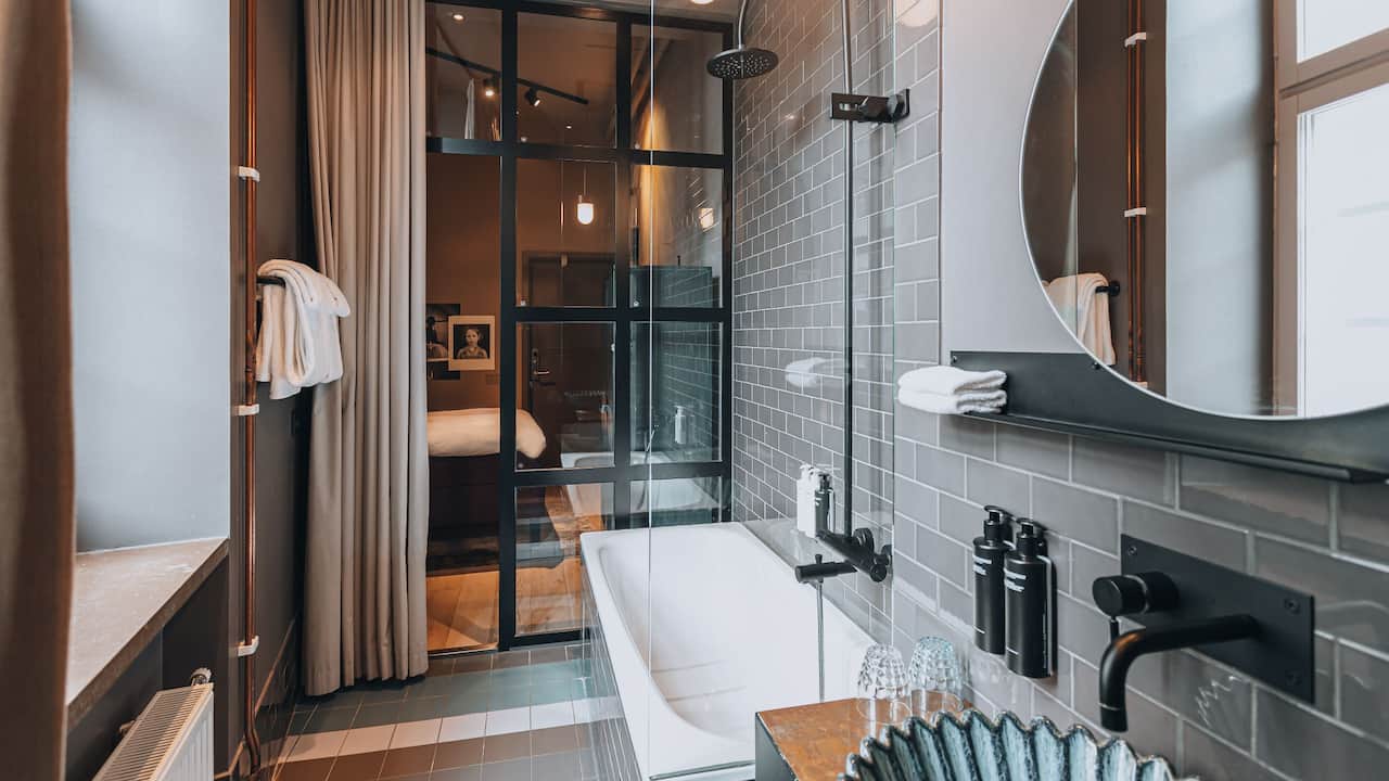 Queen Bed Deluxe Bathroom with a large mirror and a designer sink in the foreground and bathtub and shower. The hotel room showing through glass walls