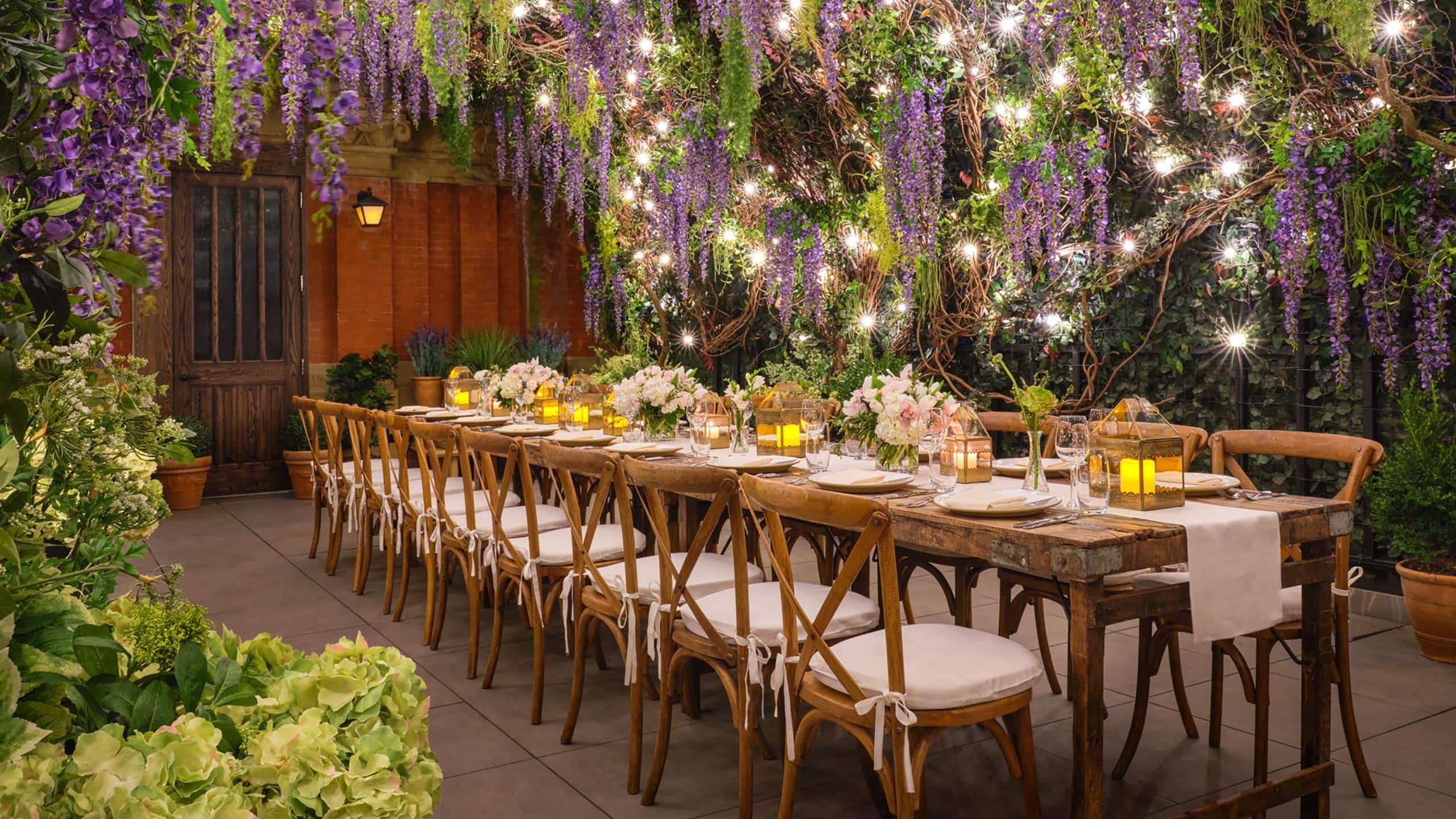 Long celebration table set with place settings and floral arrangements under vines draped with flowers and lights